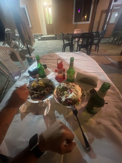 The image depicts an outdoor dining scene at night, likely in Касапите, Bulgaria, given the context. On a table covered with a white tablecloth, two plates of food are visible. One plate contains what appears to be meat with lemon slices, and the other holds a salad topped with cheese and what looks like chopped vegetables and tomatoes. Two green glass bottles of beer and two red condiment bottles are also on the table. The hands of at least two individuals are present in the frame, one holding a fork and reaching for the meat dish, and another wearing a smartwatch. In the background, outdoor seating with chairs and tables is arranged on a patio area. Lights from a building with a glass door are visible, casting shadows. The overall impression is of a casual evening meal outdoors.