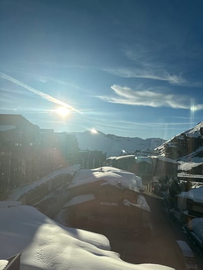 A high-angle perspective depicts a snow-covered alpine town in Saint-Martin-de-Belleville, France, under a bright blue sky with scattered cirrus clouds and a single contrail. The low sun generates substantial lens flare and long shadows across the scene. Multiple multi-story alpine buildings, featuring pitched roofs, are densely covered in snow. A prominent building in the mid-ground, identified by "BOWLING" signage on its facade, exhibits a heavy snow load on its roof. A dark, human-like figure, resembling a person on skis, is positioned on the snow-covered roof of the "BOWLING" building. A winding, partially cleared roadway traverses the urban environment, with vehicles present. Snow-covered mountain peaks are visible in the background, exhibiting sun reflections.
