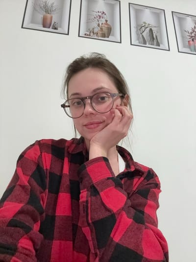 A female subject is depicted in a head-and-shoulders portrait, centrally positioned and looking directly forward. She has light brown hair and wears translucent brown-framed eyeglasses. Her right hand rests under her chin, with fingers gently supporting her jawline. She is wearing a red and black buffalo plaid flannel shirt, partially unbuttoned to reveal a white garment underneath. The background consists of a plain white wall adorned with four framed decorative images. From left to right, these images illustrate: a vase with dried vegetation and two small animal figurines; a vase with pink flowers and two small deer figurines; an arrangement of rocks and a bare tree branch; and a vase holding red flowers. The scene takes place in an indoor environment. There are no discernible visual cues within the image to confirm its location as Ercolano, Italy.