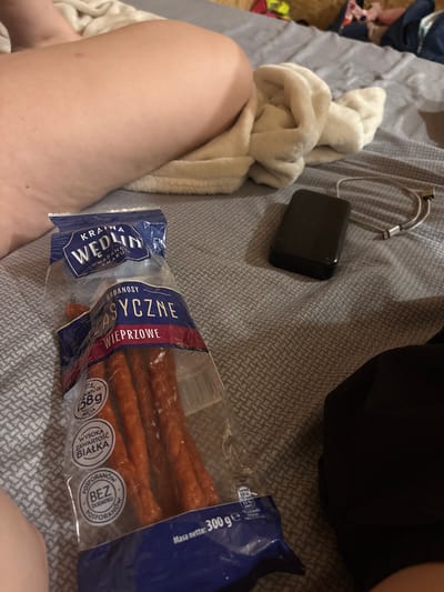 A close-up, high-angle shot shows a person's left leg resting on a bed with a grey, patterned comforter. To the right of the leg is a white fluffy blanket. In the middle of the frame is a clear plastic bag of Kabanosy Klasyczne Wieprzowe, which are pork kabanos sausages. The bag has blue and white labeling. Below the bag, a black power bank and a silver charging cable are visible on the bed. The scene appears to be indoors, likely in a bedroom. The image does not provide any specific information to determine the location in Mszczonów, Poland.
