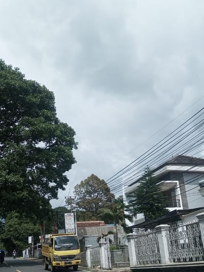 A yellow truck is parked on the side of a road, next to a white, ornate fence in front of a modern house. A large, green tree is on the left of the frame, while power lines stretch across the sky. The sky is overcast and gray. Other trees can be seen in the background, along with a building with a sign. A motorcycle passes on the road.