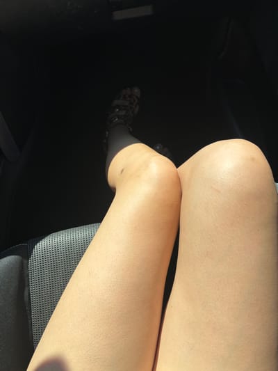 The image shows a person's legs and feet inside a vehicle, likely on the passenger side. The legs are bare and show skin texture. The person is wearing leopard print sandals with ankle straps. The interior of the vehicle is dark, suggesting it is either daytime with sunlight coming through the window or nighttime. The seat appears to be dark leather with a textured pattern. The location is not discernible from the image.