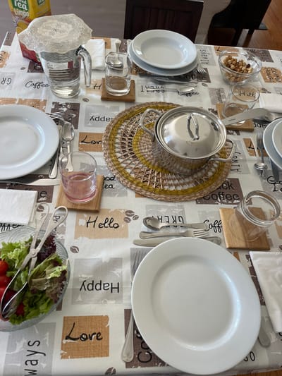 This image depicts a dining table, likely within a residence in Oliveira de Frades, Portugal, though the specific location within the city is not discernible from the visual information. The table is covered with a white tablecloth featuring repeating square patterns with words such as "Coffee," "Hello," "Happy," and "Love" alongside coffee bean motifs.

The table is set for a meal, with multiple individual place settings partially visible. Each setting includes a white circular plate and an assortment of stainless steel cutlery (knives, forks, spoons). Several clear glass tumblers are present, some resting on light-colored wooden coasters; one glass contains a pink liquid, while others appear empty or contain clear liquid. White folded paper napkins are also part of the settings.

Centrally located on the table is a round, woven placemat with yellow and natural straw tones, upon which rests a silver-colored stainless steel pot with a matching lid and two handles. To the upper left, a clear glass pitcher filled with water is covered by a white lace doily. A yellow carton of "Nestea Iced Tea Limão" is positioned behind the pitcher.

To the lower left, a clear glass bowl contains a salad of mixed green lettuce and red cherry tomatoes, with metal salad tongs partially submerged within. In the upper right, another clear glass bowl holds an assortment of nuts, including hazelnuts and almonds. No individuals are visible in the image. The background shows dark wooden furniture and light flooring, indicating an indoor setting.
