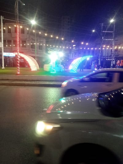 The image depicts a nighttime street scene in Majengo ward, Kenya, characterized by urban illumination and vehicular traffic. In the foreground, two light-colored vehicles are in motion on a wet asphalt road, showing blurred headlights and reflections of overhead lights on their hoods. The closest vehicle, partially visible at the bottom, displays a bright white headlight.

A central median or island separates the road from the background buildings. This median features several illuminated decorative elements: a tall pole wrapped in red string lights on the left, followed by two white, arch-like structures. To their right, a brightly lit central monument or sculpture glows predominantly green and white, with swirling elements at its apex. Adjacent to this, a blue LED-lit arch structure is visible. Several indistinct individuals are present on the median, appearing to be observing the lights; one person near the blue arch wears an orange safety vest.

In the background, multi-story buildings line the street, some with illuminated windows. Visible signage includes "FUSANI CHEMIST" and "Safcom m-pesa." Overhead, warm-toned string lights are strung across the street, contributing to the ambient illumination. Utility poles and power lines are also evident against the dark sky. The overall environment is an active urban area at night, enhanced by decorative lighting.