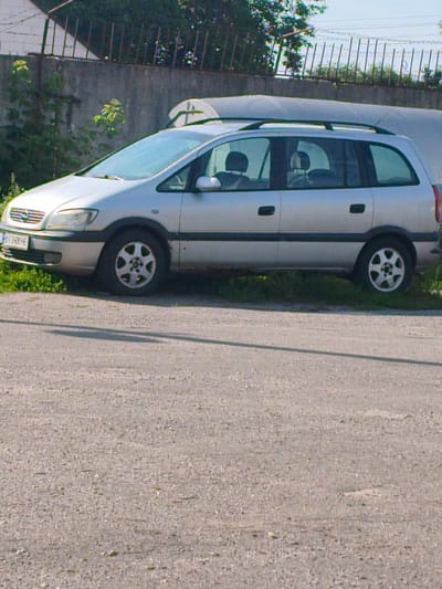 A silver minivan is parked on a paved surface next to a patch of grass. The minivan has a roof rack and tinted windows. A license plate on the front of the vehicle reads "BY 2400 EE". The background shows a concrete wall and a fence with some greenery. The scene appears to be in an outdoor setting, possibly a street or parking area in Lubny, Ukraine.