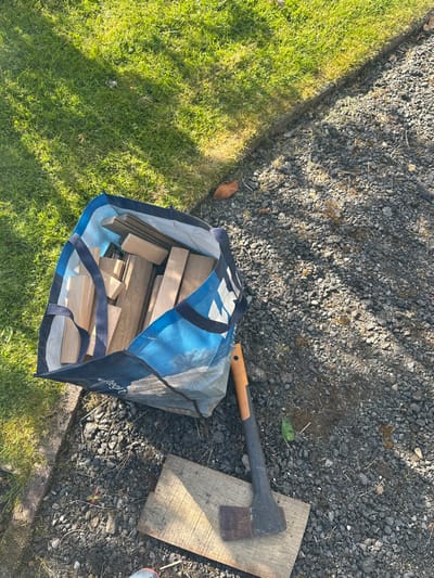 The image shows a blue and white bag filled with wooden planks, lying on a gravel path near a wooden plank and an axe with an orange handle. The path is located next to a green lawn. The location is in Cloughton, UK. The coordinates of the location are 54.369, -0.506.