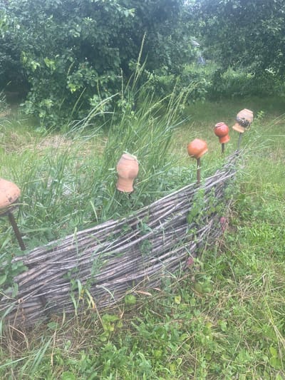 In Novyi Tahamlyk, Ukraine, a rustic, low fence constructed from intertwined branches runs horizontally through a grassy area. Several clay pots are impaled on metal stakes, protruding from the ground and serving as decorative elements along the fence. One pot, on the left, is a simple, wide-mouthed vessel. To its right, a small, rounded orange pot sits on a stake, followed by another similar orange pot. Further to the right, a more ornate, light-brown pot with decorative dark patterns rests on a stake. The background is filled with lush green foliage and tall grass, suggesting an outdoor, possibly garden or natural setting. The scene appears to be a quaint, handmade decorative arrangement in a semi-wild environment.