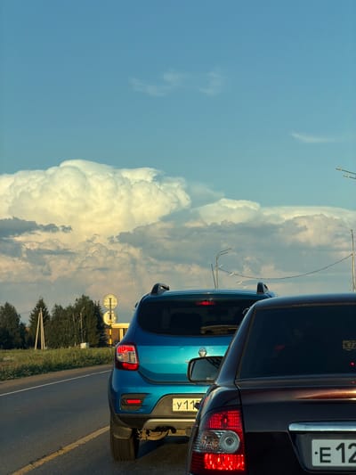 The image shows a traffic scene with two cars on a road. The sky is blue with large, dramatic clouds. The foreground car is dark brown and its rear is visible. Behind it is a bright blue car. The road appears to be asphalt with yellow lane markings. Trees and utility poles are visible on the left side of the road, along with traffic signs. The scene is likely in a semi-urban or rural area, possibly on the outskirts of Derzhavino, Russia, given the presence of the license plates. There are no people visible in the image.