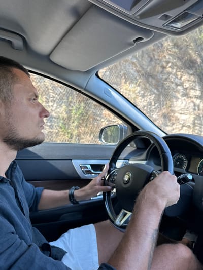 A man is driving a car on a road in Prijeradi, Montenegro. He is looking forward, with his hands on the steering wheel. The car's interior is visible, including the dashboard and steering wheel, which has a Jaguar logo. The man is wearing a dark blue shirt and white shorts. Outside the car, rocky terrain and vegetation can be seen. The image appears to be taken from inside the car, looking forward.