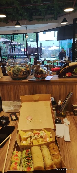 The image depicts an indoor dining area within a restaurant setting in Tumbaco, Ecuador, featuring a view of an outdoor patio. In the immediate foreground, an open cardboard take-out box rests on a wooden table. The box contains two compartments: the upper one holds nachos topped with a white sauce, what appears to be diced shrimp or chicken, and various herbs; the lower compartment contains four distinct rows of food rolls, including two varieties of sushi rolls (one with green topping and black sesame seeds, another with red and white sauces) and two types of fried or baked rolls. To the left of the box are a black smartphone, black earbuds, and a pair of wooden chopsticks. To the right, a clear glass container holds writing implements, a silver shaker, and folded white paper napkins.
Behind the table, a wooden counter features a large glass fishbowl-style container filled with wrapped items, a smaller similar container, and a decorative wooden object resembling a taco. Beyond the counter, large glass windows reveal an outdoor garden area. This exterior space contains several wooden dining tables and chairs, a wooden gazebo-like structure, green foliage, a white outdoor umbrella, and a distant white building. A single individual, wearing a dark jacket and a green cap, stands near a table in the outdoor area on the right side of the frame. The interior ceiling above the counter is adorned with artificial vines and black pendant lights. The timestamp "24/02/2026 17:37" is visible in the lower right corner.