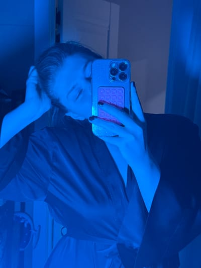 A person is holding a smartphone with a pink accessory attached to the back. The phone has multiple cameras on its back. The person's hand and part of their face are visible as they appear to be taking a selfie or looking at the phone. They are wearing a dark-colored robe or shirt. The lighting in the scene is predominantly blue, casting a strong blue hue over the entire image.