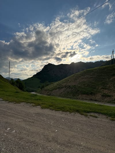 This media file depicts a scenic view of a mountain landscape with a dirt road in the foreground. The scene is dominated by the rugged slopes of a mountain covered in green grass and sparse trees. A winding dirt road, bordered by a strip of vibrant green grass, runs along the right side of the frame. In the background, towering mountains rise, their dark, rocky surfaces contrasting with the bright, cloudy sky.
The sky is a mix of blue and white, with dramatic, sun-drenched clouds creating a sense of depth and light. The sun is partially obscured by the clouds, suggesting either early morning or late afternoon light. The overall mood is serene and natural, highlighting the raw beauty of the mountainous terrain. There are visible power lines stretching across the sky, indicating the presence of some infrastructure, but they do not detract from the natural splendor.
The location is identified as Nizhnyaya Mara, Russia, which provides context for the mountainous and possibly remote setting. The image captures a moment of natural tranquility, with no people or specific activities taking place, allowing the viewer to appreciate the expansive landscape and atmospheric conditions. The textures of the dirt road, the grassy slopes, and the cloudy sky are all rendered with detail, contributing to the immersive quality of the image.