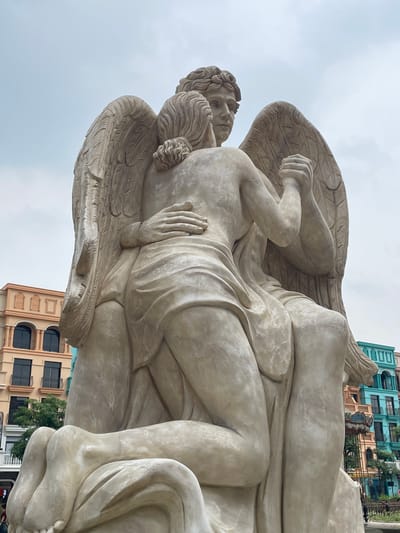 This is a medium shot, eye-level, outdoor photograph of a large, light-colored stone statue of two winged figures embracing. The statue depicts two figures with wings, one kneeling and embracing the other, who is seated. Both figures are partially draped in flowing fabric. The statue is positioned in front of a backdrop of colorful, European-style buildings and a cloudy sky. This statue is located at the Sun World complex in Nghĩa Trụ Commune, Vietnam.