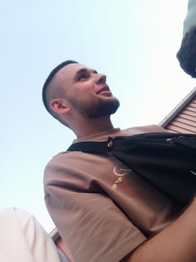 This is a close-up, low-angle shot of a young man, viewed from the chest up, with the sky as the background. He has short, dark hair with a buzz cut and a beard. He is wearing a light brown t-shirt with a circular graphic design and a black crossbody bag with a Nike logo visible. He is smiling and looking to his right. The setting appears to be outdoors during the day, with a bright, clear sky. In the background, there is a reddish-brown building with horizontal siding, possibly a barn or shed. The lighting suggests it could be late afternoon or early evening. The overall mood is casual and relaxed.