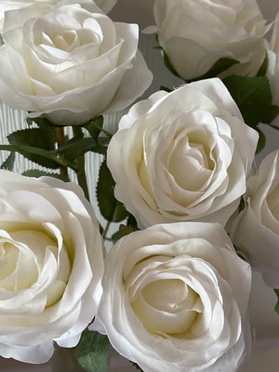 The image is a close-up, eye-level, medium shot of a bouquet of artificial white roses. The roses are in full bloom, with multiple layers of petals. Some of the roses are slightly out of focus, creating a depth-of-field effect. Green artificial leaves are visible interspersed among the flowers, and the stems are a light brown color. The background is a light gray color with a subtle vertical striped pattern. There are no people or animals visible in the image, and no specific location in Bielefeld, Germany, is identifiable. The scene is static and appears to be an indoor setting.