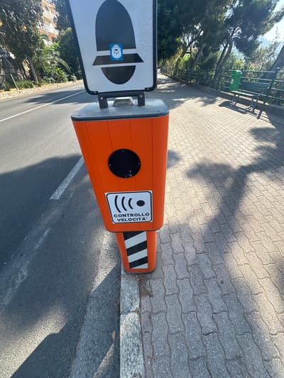 This is a close-up, eye-level, medium shot of an orange speed camera on a paved sidewalk next to a road. The camera is a bright orange, hexagonal pillar with a black circle at the top and a sign with the Italian words "CONTROLLO VELOCITA'" (speed control) below it, accompanied by a radio wave symbol. A blue sticker with a cartoon character wearing a hat is adhered to the top of the camera. The road is asphalt with a white line marking the lane. The sidewalk is made of interlocking gray pavers. In the background, there are trees, a green bench, and a green trash can. The scene is illuminated by sunlight, casting shadows on the sidewalk. The environment appears to be outdoors in a town or city setting.