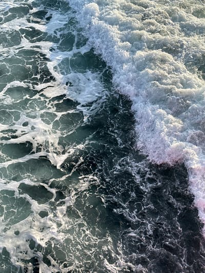 The image displays a close-up, downward view of turbulent ocean water in the city of Palaiopoli, Greece. A prominent breaking wave, characterized by dense, bright white foam, extends diagonally across the upper right portion of the frame. To the left of this breaking wave, the water surface exhibits a dark teal-green hue interspersed with an intricate network of white foam patterns, indicating agitated water movement and aeration. No human subjects or discernible man-made objects are present in the scene; the composition focuses exclusively on the dynamic interaction of water and foam.