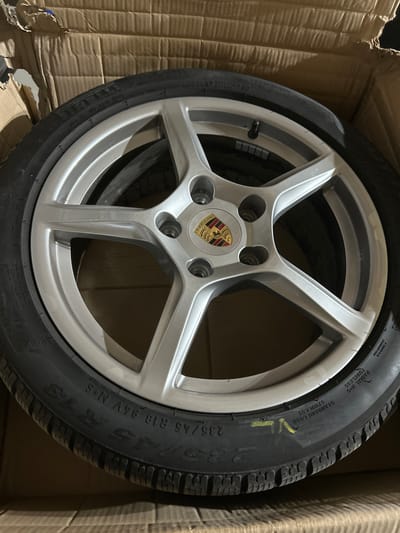 A silver Porsche alloy wheel with a tire is pictured inside a cardboard box. The wheel has five spokes and a Porsche crest in the center. The tire has lettering on its sidewall, which is partially obscured. The background is the inside of the cardboard box, which is brown and textured. The image was taken in the city of Djerman, Bulgaria. There are no people or actions depicted in the image.