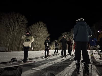 At night, a group of people are standing on a snowy surface, possibly a ski slope, illuminated by artificial lights. Most are wearing ski gear, including helmets and skis, and are holding ski poles. The foreground includes a person with a snowboard. A wooded area is visible in the background, with bare trees against the dark sky. The scene suggests a nighttime skiing or snowboarding event.