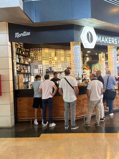 The image shows the exterior of a cafe with the sign "Morettino" and a coffee bean logo. Several people are standing in front of a counter, appearing to be customers or staff. The cafe has a tiled wall with a geometric pattern and wooden accents. The lighting fixtures are visible hanging from the ceiling. The floor is tiled with a marble pattern. The overall scene suggests a commercial establishment, possibly a coffee shop or cafe, within an indoor public space. The provided hint mentions the city of Carini, Italy, which places this scene in that specific location.