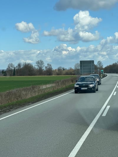 The scene depicts a rural road under a partly cloudy blue sky, presumably within the vicinity of Kirchdorf am Inn, Germany. The asphalt road features two visible lanes, separated by a dashed white line, with a solid white line marking the right shoulder from the perspective of the camera. The road curves to the right in the distance.

Three vehicles are present on the road. In the foreground, traveling towards the camera, is a black BMW sedan with its headlights illuminated. Its German license plate reads "AB CH 106". Directly behind the BMW is a second dark-colored sedan, partially obscured. Further down the road, near the curve, a third vehicle, possibly a light-colored truck, is visible.

To the left of the road, a wide, vibrant green field extends, bordered by a line of dry, brown shrubbery or weeds along the roadside. Beyond the field, a line of deciduous trees, largely bare, stands against the horizon. To the right of the road, a dense treeline runs parallel to the asphalt. A rectangular, grey road sign with its back facing the camera is positioned on the right side of the road, preceding the curve. The sky above is characterized by scattered white cumulus clouds. No individuals are visible in the image. The photograph appears to be taken from a moving vehicle.
