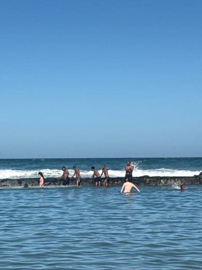 The image depicts a scene at Muizenberg Beach in South Africa. A long, low concrete breakwater stretches into the ocean. Several people are positioned along the breakwater, with some leaning over the edge, as if preparing to jump into the water. The ocean is visible on one side of the breakwater, showing the waves and water surface, and the sky is visible on the other. The day seems clear, with no cloud cover.