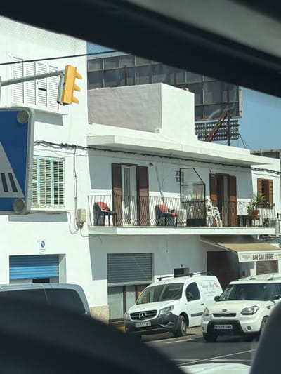 This is a vertical, medium-shot, outdoor photograph taken from inside a vehicle, looking out. The scene is a street in what appears to be a Mediterranean town, possibly Santa Eulària des Riu, Spain, given the context. The building in the foreground is a white, multi-story structure with a modern facade, featuring several windows with dark brown shutters and balconies with metal railings. Some of the windows are covered by metal grates. A bright yellow traffic light with three lights is visible on the left side of the image, mounted on a pole.
In the background, a slightly older building with a light gray, textured facade can be seen. It has a section with large, square windows. Further to the right, a newer, white van with a black stripe and the text "VANSISTEM" is parked. Another white van, possibly a Kia Soul, is parked next to it, with a license plate that reads "S329 GMR". A sign for "BAR CAÑ NEGRE" is also visible on the building.
The time of day appears to be daytime, likely late morning or early afternoon, with clear, bright sunlight casting shadows. The weather seems to be warm and sunny, typical of a Mediterranean climate. The perspective suggests the photographer is in motion, possibly driving through the town. No people are clearly visible. The overall impression is one of a typical urban street scene in a sunny European location.