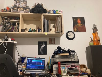 The image captures a detailed view of a personal home office or studio space in Kokhav Ya'ir Tzur Yigal, Israel, characterized by a mix of functional electronics and decorative items.
Above a light-colored wooden shelving unit, several Funko Pop figures in their boxes are stacked. A large, dark, plush gorilla toy sits beside them, next to a small model sailing ship. The shelving unit itself holds an assortment of items: the left side of the top shelf contains books and various figurines, including a white rabbit. The right side holds what appears to be a stack of records. The bottom shelf on the left has more books and decorative objects, including a small religious figurine and a pot. A yellow smiley face sticker is attached to the front edge of this shelf.
On the white wall to the right of the shelving, a printed image of a person wearing a crown and holding a scepter, styled like a classical portrait, is displayed. Below it, a round black wall clock indicates the time as approximately 4:45. Further to the right, an orange vase and an orange-tinted bottle sit on a dark surface, accompanied by a small green candle. Below the shelving on the wall is a black and white poster of a person playing a trumpet.
The desk area below is laden with electronic equipment and personal items. A silver laptop, open and displaying what appears to be audio editing software with prominent blue waveforms, is central. To its right, a black microphone rests on a stack of audio equipment, which includes a silver turntable and an amplifier or receiver. A bright red pepper grinder stands on the desk, surrounded by various cables, small bottles, and other small accessories. Part of a dark office chair is visible on the far left, and a large, dark speaker cabinet stands on the right side of the frame. The scene suggests a lived-in environment focused on music production, listening, or general computing. No clearly legible text is visible other than the time on the clock. The lighting indicates an indoor setting, likely under artificial light.