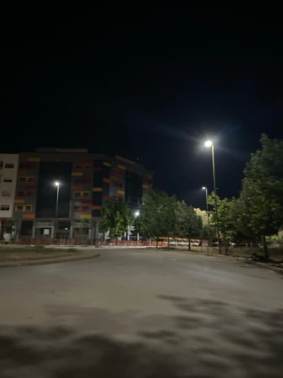 The image depicts a street scene at night in Dar Oum Soltane, Morocco. A multi-story building with brightly colored window frames faces the street. Trees line the road. Streetlights illuminate the scene. The road is wet. The sky is dark. There are no people visible.