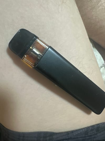 A black vape pen with a visible amber liquid cartridge rests on a person's leg, which is covered by dark grey fabric at the bottom of the frame. The background is a light, textured surface. There is no indication of the city of Corlăteni, Moldova.