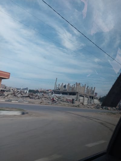 The image depicts a severely damaged urban street scene in a-Zawayda, Palestinian Territory, observed from a vehicle. In the foreground, a grey asphalt road, possibly an intersection, curves from the lower left towards the center and right. Reflections are visible on the vehicle's window in the upper right. The midground is dominated by extensive piles of concrete rubble and the skeletal remains of multiple multi-story concrete buildings. One large structure on the right side of the midground appears partially collapsed, with exposed rebar, broken concrete columns, and debris scattered around its base. Further back, more completely leveled structures form a landscape of debris. A partial, orange-brown structure is visible on the far left. The background features a light blue sky with scattered cirrus clouds and multiple dark power lines crossing horizontally. No individuals or active subjects are visible within the scene.