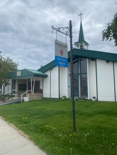 The image shows the exterior of Christ Church Cathedral, located in Whitehorse, Canada. The building is a modest structure with white vertical siding and a green roof. A steeple with a cross rises from the roof, and a bell is visible within the steeple. A sign in front of the building reads "THE ANGLICAN CHURCH WELCOMES YOU" and "Christ Church Cathedral Whitehorse." The church has large stained-glass windows on the side. The grounds are covered in grass with scattered dandelions and some potted flowers near the entrance. A sidewalk runs along the left side of the image. The sky is overcast. There are no people visible in the image.