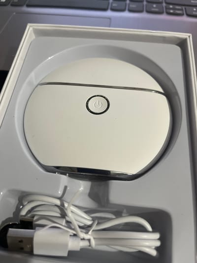 The image shows a white, oval-shaped electronic device with a silver trim and a circular power button on its surface, resting in a white plastic packaging. A white USB charging cable is coiled beside the device. The device appears to be new, as it is presented in its original packaging, suggesting it is a product for sale or recently purchased. The background includes a portion of a laptop keyboard, indicating the item is likely being photographed in a home or office setting. There is no visible text in the image. The lighting suggests an indoor environment, possibly during the daytime. The overall scene is neutral and focused on showcasing the product.