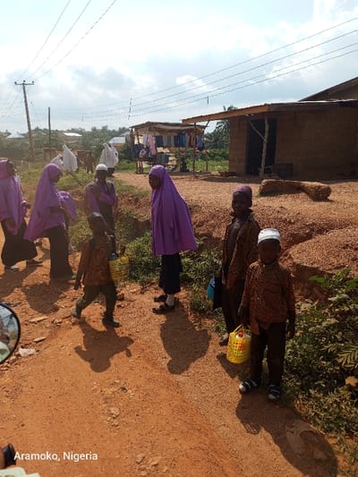 The image shows a group of people, mostly children, in a rural street in Ilobu, Nigeria. The street is unpaved and dusty. The children are carrying baskets, suggesting they might be returning from a market or school. The background shows a modest house with a thatched roof, a utility pole with power lines, and a few trees. The scene seems to be in a rural part of the town, with a lot of dust and dirt. There are six children visible in the scene: three girls and three boys. The girls are wearing hijab, and the boys are wearing caps. Some of the children are carrying baskets in their hands, which are likely filled with groceries or other items. One child is standing on the side of the street looking at the camera. The children are dressed in simple clothing, and the overall scene suggests a rural and modest setting.