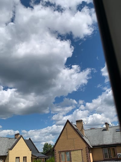 The image depicts a bright, cloudy sky over the rooftops of residential buildings in Martem'yanovo, Russia. The day appears to be warm and sunny, with large, fluffy white clouds scattered across a vivid blue sky, interspersed with some darker, grayer clouds that suggest potential for rain. The perspective is from a high vantage point, possibly a window, looking upwards and slightly to the side.
The foreground is partially obscured by a dark, horizontal frame, suggesting it is taken from inside a building or structure. Below the sky, the angled roofs of several houses are visible. These houses have dark gray shingled roofs and walls of a light brown or tan brick. Some architectural details, like decorative brickwork and multiple windows, are discernible on the buildings. Chimneys with satellite dishes are present on some of the roofs, indicating modern living. A small air conditioning unit is visible on one of the upper roofs. Trees can be seen in the background, suggesting a suburban or semi-rural setting.
There is no discernible activity or event taking place, it is a still scene. The time of day is likely midday or early afternoon, given the brightness of the sky and the shadows cast by the clouds and buildings. The overall mood is serene and peaceful, characterized by the interplay of sunlight and clouds. No text is visible in the image.