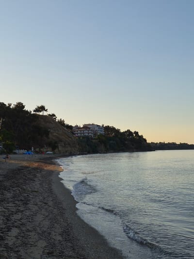The image depicts a serene beach scene at sunset in Nea Kallikratia, Greece. The foreground is dominated by a sandy beach with scattered debris and small stones, transitioning into the gentle, calm waters of the sea. The water displays subtle ripples and reflects the soft hues of the sky.
Along the shoreline, the land rises to form a vegetated cliff. Scattered on the beach are two blue and one white umbrella, suggesting a recreational area. A few small figures are present on the beach, one appearing to be seated on a bench and another standing near the water's edge.
Further back, the cliff face is covered with trees and shrubs, with a prominent line of pine trees at the crest. A multi-story hotel or apartment building, with white walls and balconies, is visible atop the cliff, overlooking the sea. The sky above is a clear gradient of pale blue to soft yellow, indicating the late hour of the day. The overall atmosphere is peaceful and tranquil.