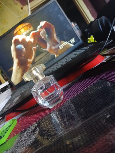 This media file captures a close-up, angled shot of a clear glass perfume bottle with a faceted cap, resting on a surface adorned with various items. The bottle itself appears to be a luxury fragrance, with "SHINE SONN" visible on a label. The immediate background is cluttered, featuring what looks like a framed image or artwork with a person depicted, and other objects with textures ranging from smooth to woven. A red, a black, and a purple textured item are prominent behind the perfume bottle. The lighting suggests an indoor setting, possibly a desk or shelf, with no clear indication of time of day or weather. The overall impression is one of personal belongings arranged in a somewhat informal setting. There are no clear cues about a specific activity or event. The location context of Chutove, Ukraine, is provided but not visually evident within the frame.