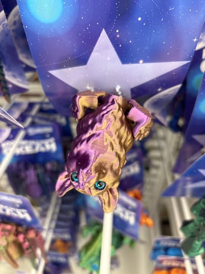 A 3D printed figurine of a small, golden-purple creature with bright blue eyes is the primary subject. It is perched on a blue and white patterned surface featuring stars and a large white star, suggesting a space or fantasy theme. The figurine is angled slightly upwards, with its front paws resting on the surface. In the background, other similar figurines in various colors like green, blue, and orange are visible, hanging in what appears to be a retail display. These are also in plastic packaging with blue and white labeling. The overall scene is brightly lit, indicative of an indoor setting like a store. No people are visible. The image is taken from a close-up perspective, focusing on the central figurine, with the background elements blurred, creating a shallow depth of field. The background suggests a store aisle, possibly a toy or novelty store. There is no visible text that can be clearly deciphered.