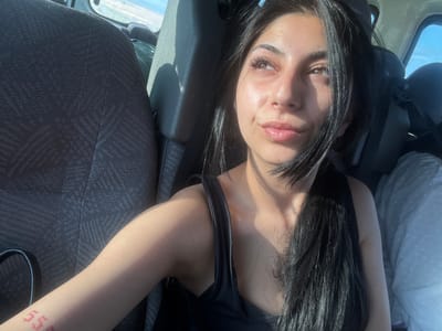 The image shows a woman with long, dark hair wearing a black tank top. She is indoors, possibly in a car, as a seat and what appears to be a seatbelt are visible. Part of the car's interior is gray. A tattoo is visible on her right arm. Outside the window, a sliver of light suggests an outdoor environment, and a body of water is possibly visible. The image's angle suggests it was taken from above, looking down at the subject. The location is unknown and cannot be identified from the given information. It is only provided to be within the city of Municipal Unit of Nikea, Greece.