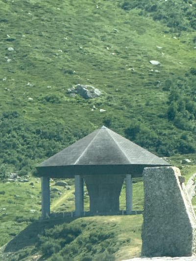 The image depicts a hexagonal, grey, multi-level structure with a dark roof, supported by four cylindrical pillars. The structure is situated on a grassy hillside, with a path winding behind it. To the right of the structure, a large, rough-hewn stone monument stands. The background is a lush, green, mountainous landscape with scattered rocks and trees. The scene appears to be a public space or a monument, likely in a rural or mountainous area. This specific structure is the Walter Gropius Memorial, a tribute to the renowned architect, located in the Swiss municipality of Hospental, within the canton of Uri. The memorial was designed by Gropius himself and was unveiled in 1967.