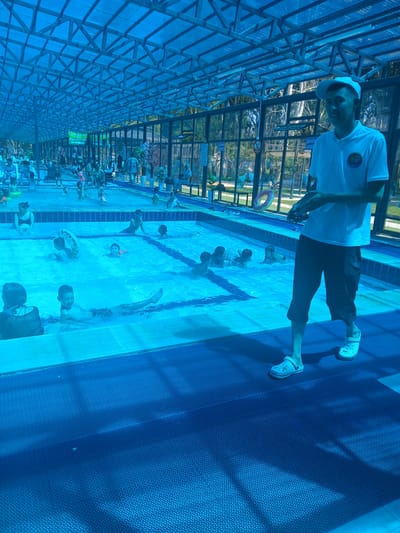 The image depicts an indoor swimming pool area, likely a public facility, with a blue, translucent roof structure. The pool is filled with water and has several people, primarily children, swimming and playing in it. Some adults are also present, supervising or relaxing. A young man wearing a white t-shirt, dark shorts, and a white cap is standing on the poolside, holding what appears to be a remote control or phone. He is facing towards the pool, suggesting he might be an attendant or lifeguard. The area is clean and well-maintained, with blue rubber mats on the poolside. The overall impression is of a recreational and family-friendly environment. The location is identified as Boz Bulun, Kyrgyzstan.
