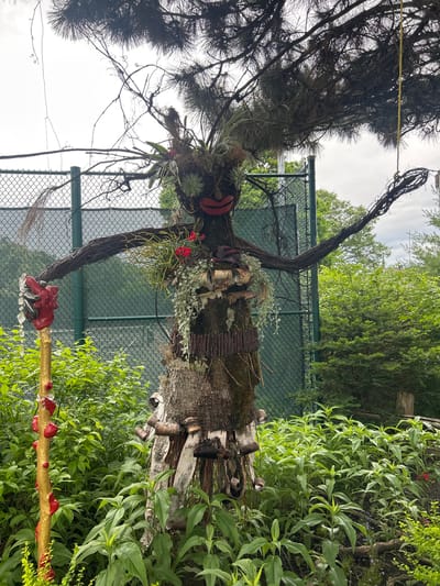 An outdoor, full-shot image captures a whimsical, human-like effigy crafted from natural materials, situated in a garden setting. The effigy, resembling a tree-like figure, is adorned with moss, branches, red lips, and various plant elements, including small red flowers and hanging greenery. Its arms, made of twisted branches, are outstretched, with one hand holding a golden staff topped with a red, sculpted hand and clear, faceted beads. The effigy stands amidst lush green foliage and is backed by a green chain-link fence and a dense backdrop of trees. The sky above is overcast. The scene is located in the city of Mount Royal, Canada.