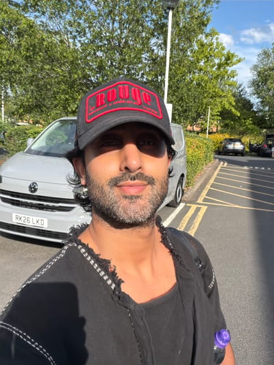 A man is captured in a selfie taken outdoors. He is wearing a black baseball cap with red lettering and a black t-shirt that has a distressed, frayed hemline with white stitching. He has facial hair and appears to be looking towards the camera with a neutral expression.
In the background, a light grey van is parked. The van has a license plate that reads "RK26 LKD". To the left of the van, trees with lush green foliage are visible, and beyond them, a bright blue sky with scattered clouds suggests it is daytime and the weather is pleasant. The ground is paved, appearing to be a parking lot with yellow lines marking parking bays. In the distance, other parked cars and a hint of yellow flowers can be seen, possibly indicating a roadside or a commercial area. The scene is illuminated by bright sunlight, casting shadows. The overall impression is a casual, everyday moment captured outdoors on a sunny day.
Visible text on the cap reads "ROUGJ" in large red letters, with smaller text below that is partially obscured but appears to include "RESTAURANT" and "APRES-SKI".