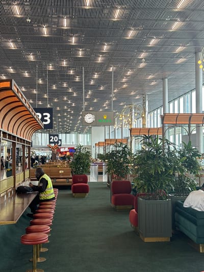 This image captures a wide interior view of a passenger terminal lounge or waiting area within Charles de Gaulle Airport (CDG) in Roissy-en-France, France. The area features a high ceiling constructed from a grid of grey metallic slats, interspersed with numerous active recessed light fixtures. The floor is covered with a dark green carpet throughout the visible space.
On the left, a long, dark wooden counter with a brass edge is fronted by a row of red, circular upholstered stools, each with a brass pedestal base. A curved, arched canopy with a light-colored, ribbed wooden interior extends above this counter. A male individual, wearing a high-visibility yellow-green vest over a dark long-sleeved shirt and dark trousers, is seated at one of these stools. He is facing right, looking down at a dark object (likely a mobile device) on the counter. A black backpack rests on the counter beside him.
The central and midground areas are furnished with various seating arrangements and potted plants. Several dark green, broad-leafed plants are contained within dark grey, slatted planters with brass bases. Multiple red upholstered armchairs, each with a brass base, are strategically placed, some featuring a "Paul..." emblem on their backrests. To the far right, a segment of a dark green upholstered sofa or bench with a brass base is visible, occupied by a person whose torso and right arm (wearing a white shirt) are discernible. A decorative light fixture featuring multiple small, spherical yellow lights on thin, curved, brass-colored stems stands in the midground.
Suspended from the ceiling are black square signs displaying white numbers "23" and "20," indicating gate or zone numbers. Adjacent to these, a rectangular sign unit exhibits an analog clock face on its left side and the green "ROLEX" logo with its crown emblem on the right side. In the background, an open concourse is visible, populated by multiple individuals walking, and a "RELAY" retail sign is identifiable. The entire right side of the terminal features large, multi-pane windows with vertical supports, allowing natural light to illuminate the space.