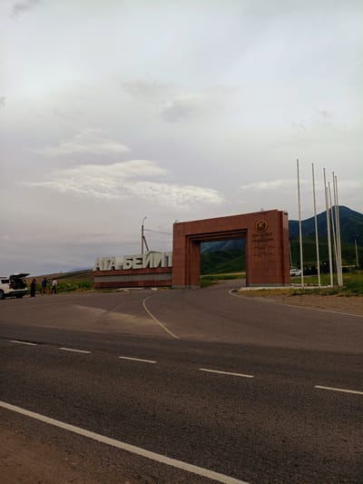 The image depicts the entrance to "ATA-BEYIT," a memorial complex located in the city of Arashan, Kyrgyzstan. A large, reddish-brown stone archway stands prominently, with the words "ATA-BEYIT" inscribed in large, white letters on a separate monument to the left. The archway itself has inscriptions and a circular emblem on its facade. Several tall, slender flagpoles stand to the right of the arch. In the background, rolling green hills and mountains are visible under a cloudy sky. In the foreground, a paved road curves towards the archway. A white SUV with its trunk open is parked to the left of the road, near the monument. Three individuals are standing by the SUV, and one appears to be holding a long pole, possibly for photography or videography. The overall scene suggests a place of historical or memorial significance, set within a natural landscape.