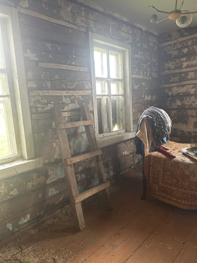 The image depicts the interior of a dilapidated structure, potentially located in Студзёнкаўскі сельскі Савет, Belarus, although this cannot be definitively confirmed from the image alone. The scene is characterized by decay and disrepair. Walls exhibit peeling paint and exposed wooden planks. A ladder leans against a wall beneath a window, possibly for accessing higher areas. A stack of clothes is placed on top of a chair near the window, adjacent to a couch. The floor is covered in debris. A light fixture hangs from the ceiling. Overall, the scene suggests abandonment and neglect.
