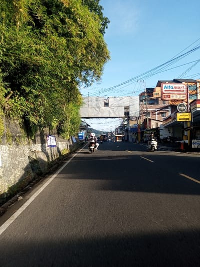 This image captures a daytime street scene in the city of Khouathineung, Laos, under a clear blue sky. The road is asphalt, marked by a solid white line on the left shoulder and yellow dashed lines separating two lanes of traffic. On the left, a low stone retaining wall supports a dense growth of green foliage, including bamboo, which overhangs the road. Several posters are affixed to this wall. Direct sunlight casts distinct shadows across the left lane of the road, suggesting morning or late afternoon.
Multiple motorcycles or scooters are present on the road, traveling away from the viewer. In the foreground, a single rider on a scooter, wearing a helmet, is in the right lane. Further down the road in the left lane, another scooter carries a rider with a white helmet and a passenger. Additional indistinct vehicles are visible in the distance.
A pedestrian overpass, constructed from what appears to be metal covered with sheeting and featuring small window-like openings, spans directly above the road. On the right side of the street, a series of multi-story buildings house various businesses. A prominent red and white "Alfamart" sign is visible on one building, accompanied by a smaller black "bean!" sign and a yellow sign with partial text below it. Numerous overhead power and communication lines are strung across the street. In the far distance, hills or mountains are visible under the clear sky.