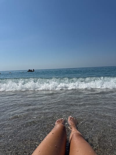The image captures a serene beach scene in Finale Ligure, Italy. The viewpoint is low, focused on the legs and feet of a person partially submerged in the clear, shallow water. The water laps gently at the shore, revealing a pebble-strewn seabed. In the distance, the tranquil blue sea stretches to the horizon under a clear sky, with a few people visible in a boat further out. The overall impression is one of peaceful relaxation on a sunny day at the coast.