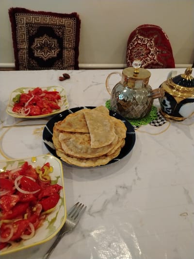 The image depicts a table set for a meal, likely in a domestic setting in Shovot, Uzbekistan. On a marble-patterned tablecloth, two square dishes filled with a chopped tomato and onion salad are visible. A stack of flat, golden-brown fried pastries rests on a dark blue plate with a decorative rim. To the right, two ornate teapots are present; one is a clear glass teapot with a gold lid, and the other is a black and gold teapot with a traditional design. Two decorative cushions, one maroon with intricate geometric patterns and another red with floral motifs, are placed behind the table. A silver fork rests on the table near one of the salad dishes. The lighting appears to be indoor ambient light. The overall scene suggests a casual meal or gathering.