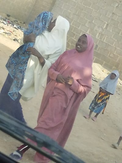 Four women, possibly a family, are gathered in an outdoor setting with a brick wall in the background. Two women are close, one embracing the other. A third woman in a pink robe is standing and smiling. A child in a hijab stands in the background wearing a patterned dress and pink shoes. The women are wearing traditional Muslim clothing. The bottom portion of the image is a vehicle windshield and frame.